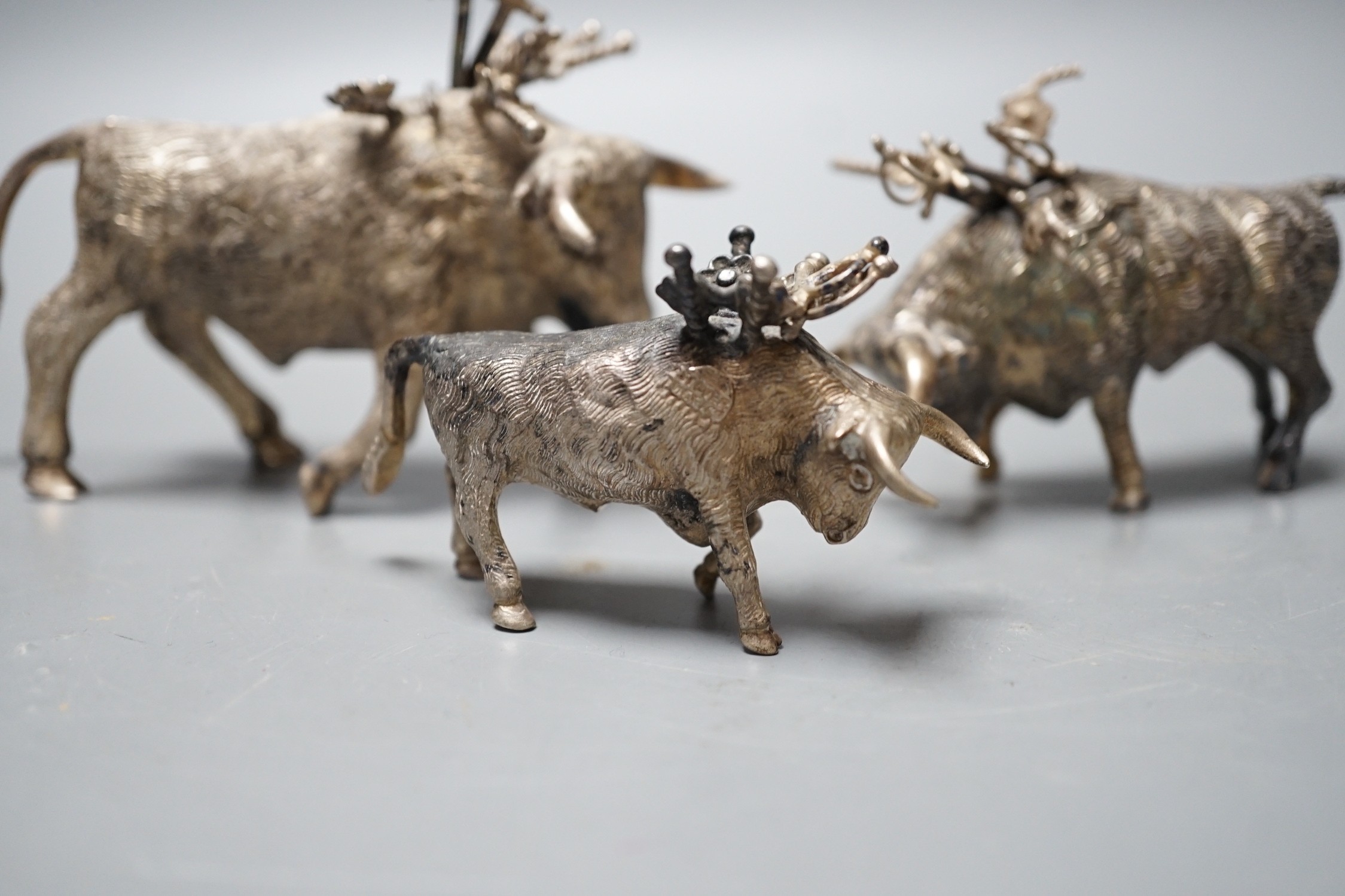 Three miniature white metal model cocktail stick holders? each modelled as a bull with sword shaped sticks, two stamped sterling 925, largest length 12.3cm.